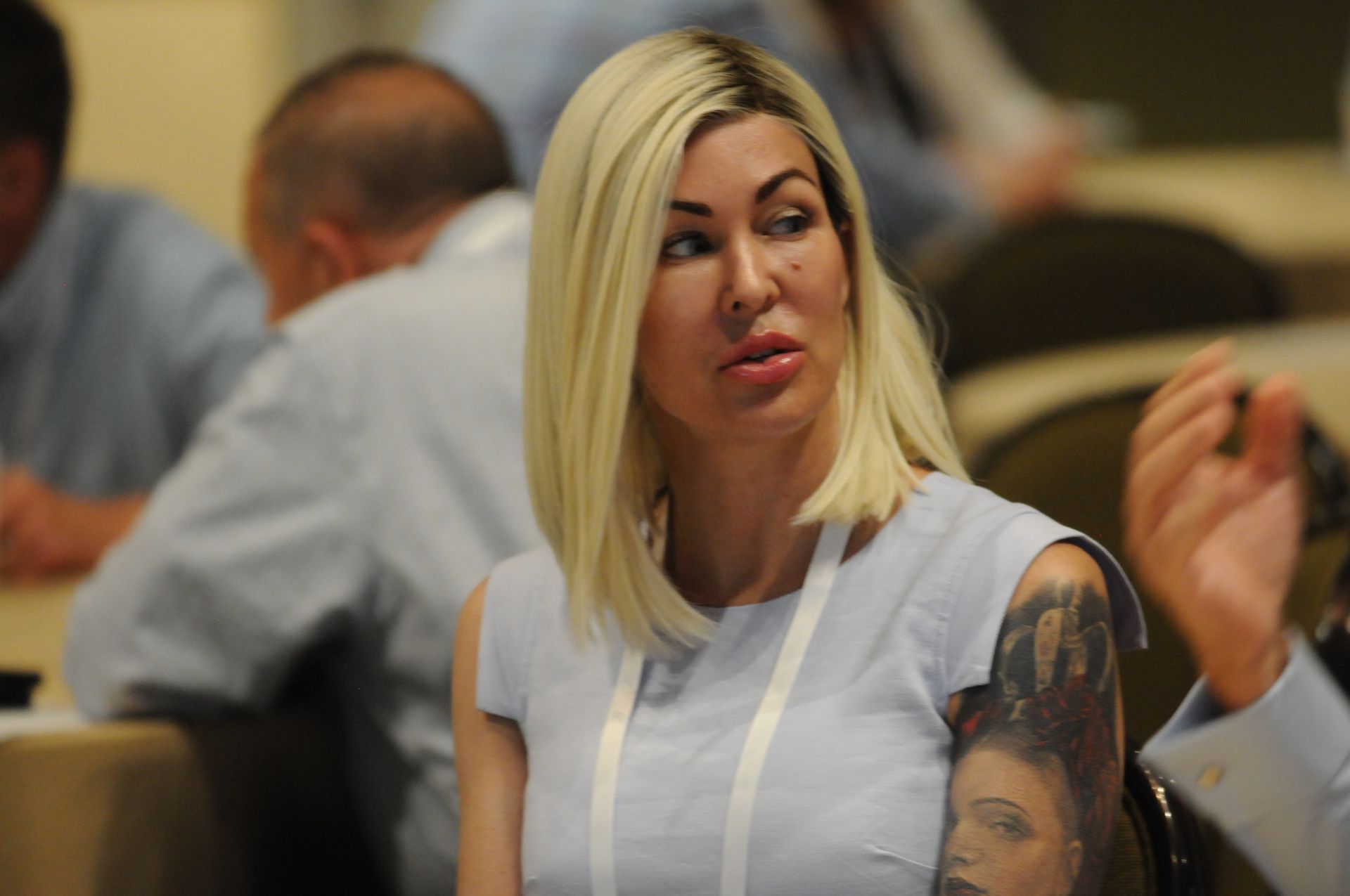 Woman with blonde hair, tattooed arm, light blue dress, seated at a conference, looking thoughtful.