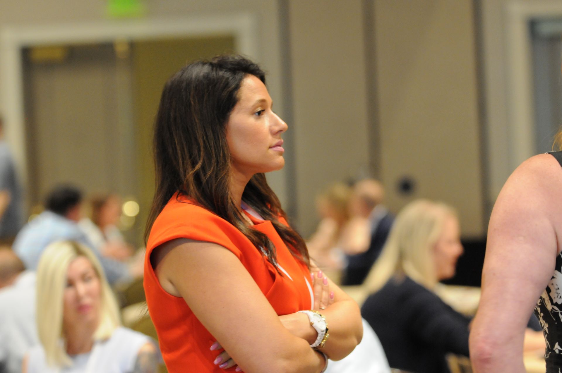Woman in orange top with arms crossed, at a conference, looking focused.