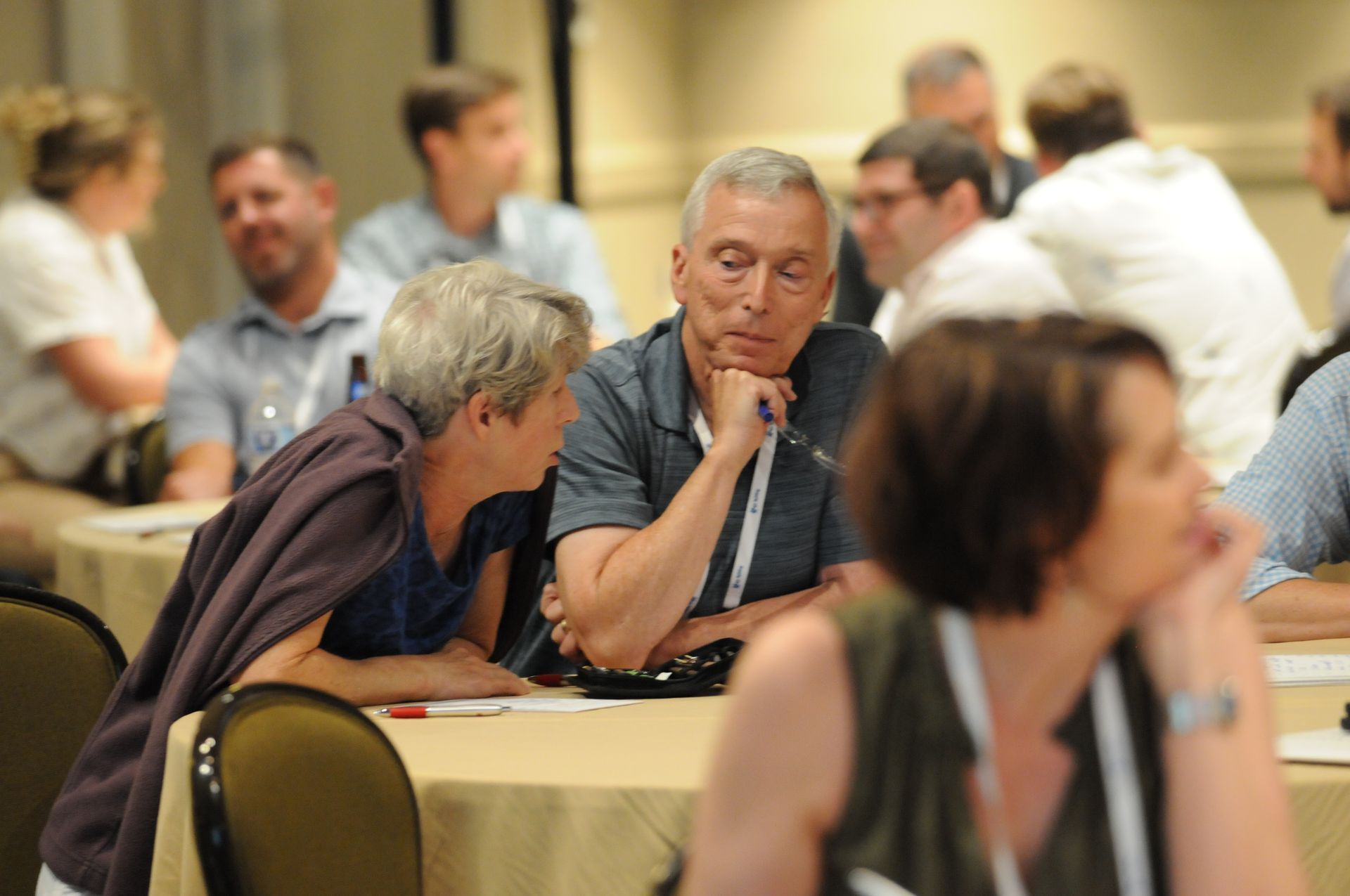 Attendees at a conference, talking at tables. A woman leans in to talk with a man. Others are in the background.