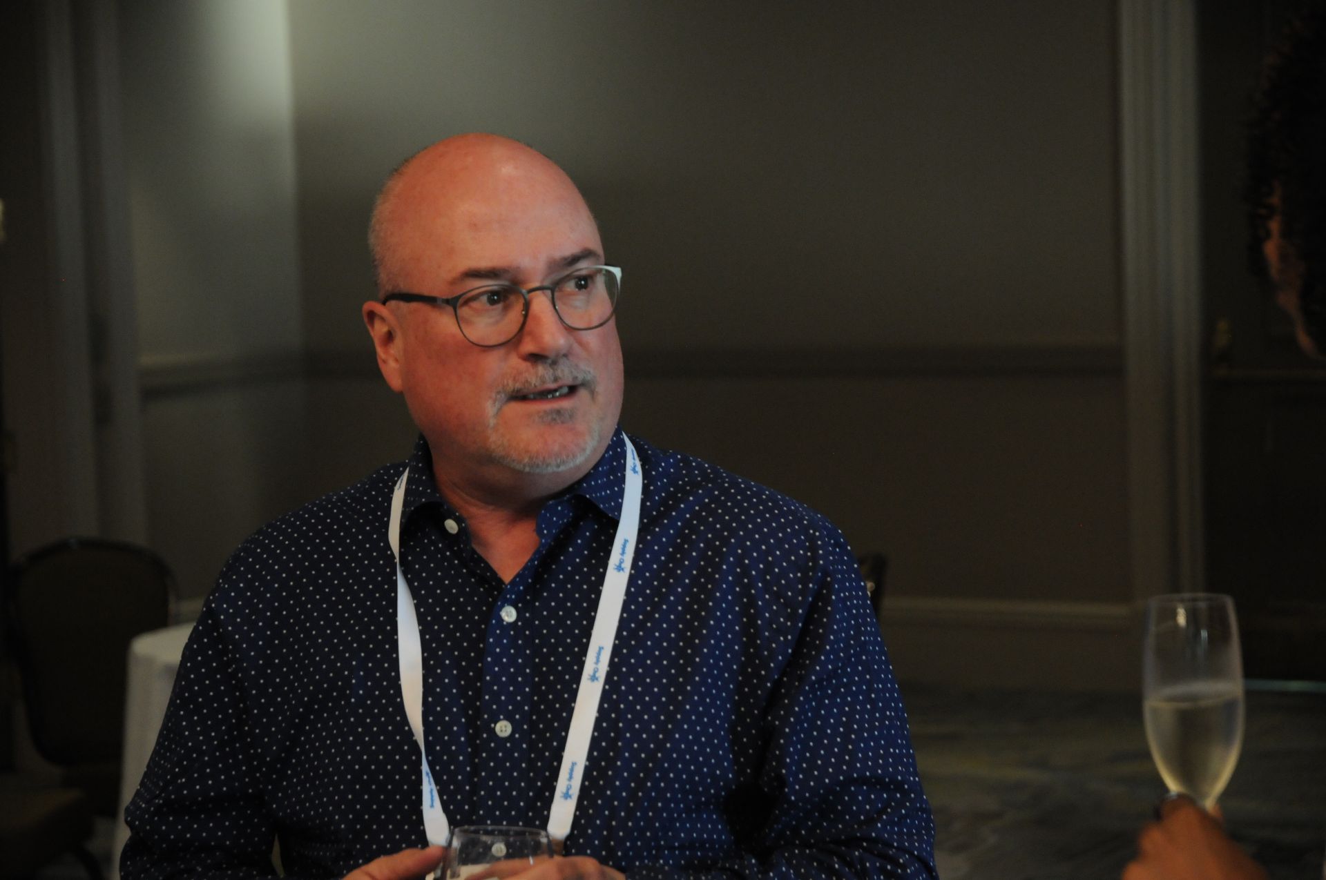 Bald man wearing glasses and blue polka-dot shirt, looking right, white lanyard around neck, indoors, possible conference.