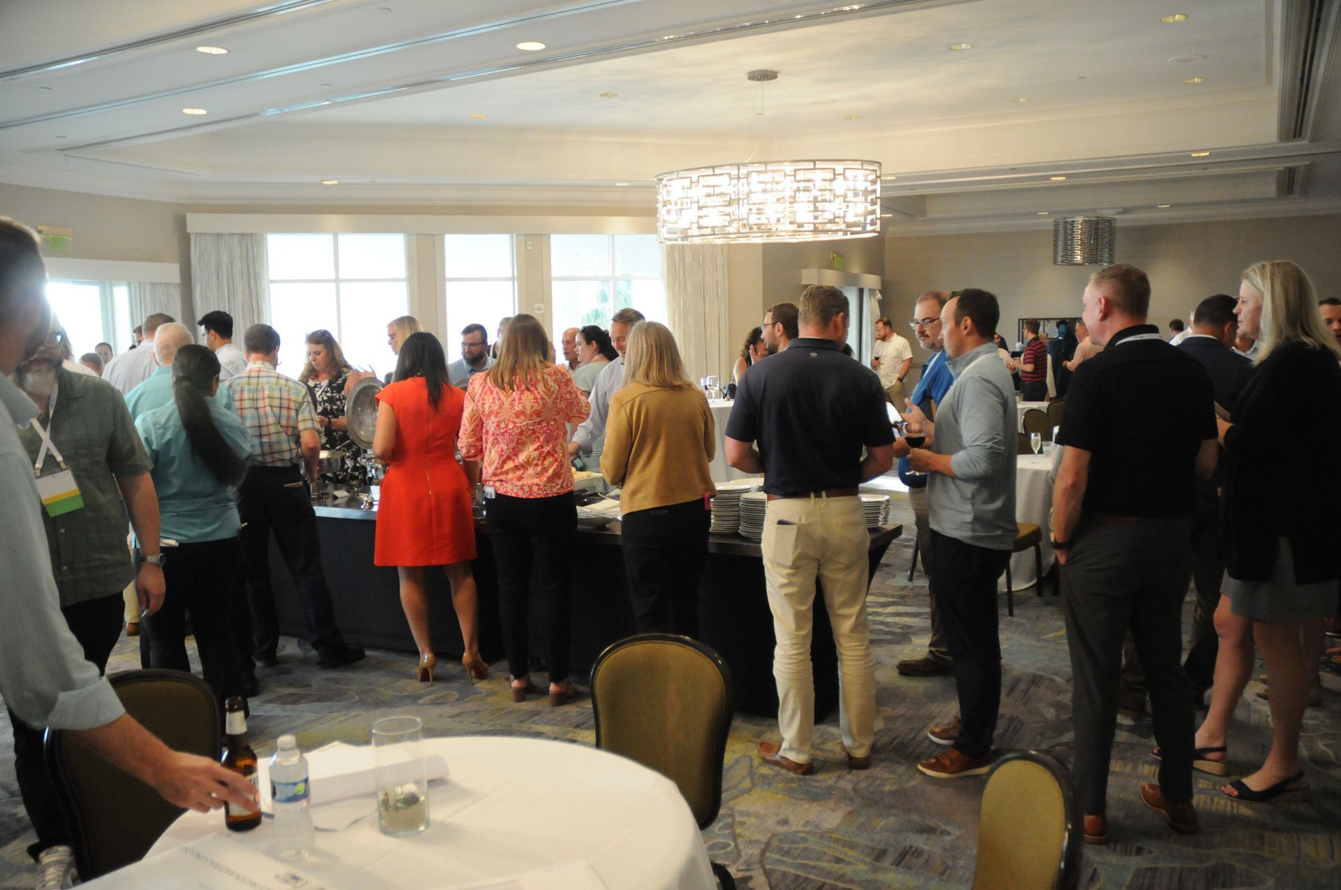 People mingling at an event, some near a buffet table, in a well-lit room with windows and round tables.