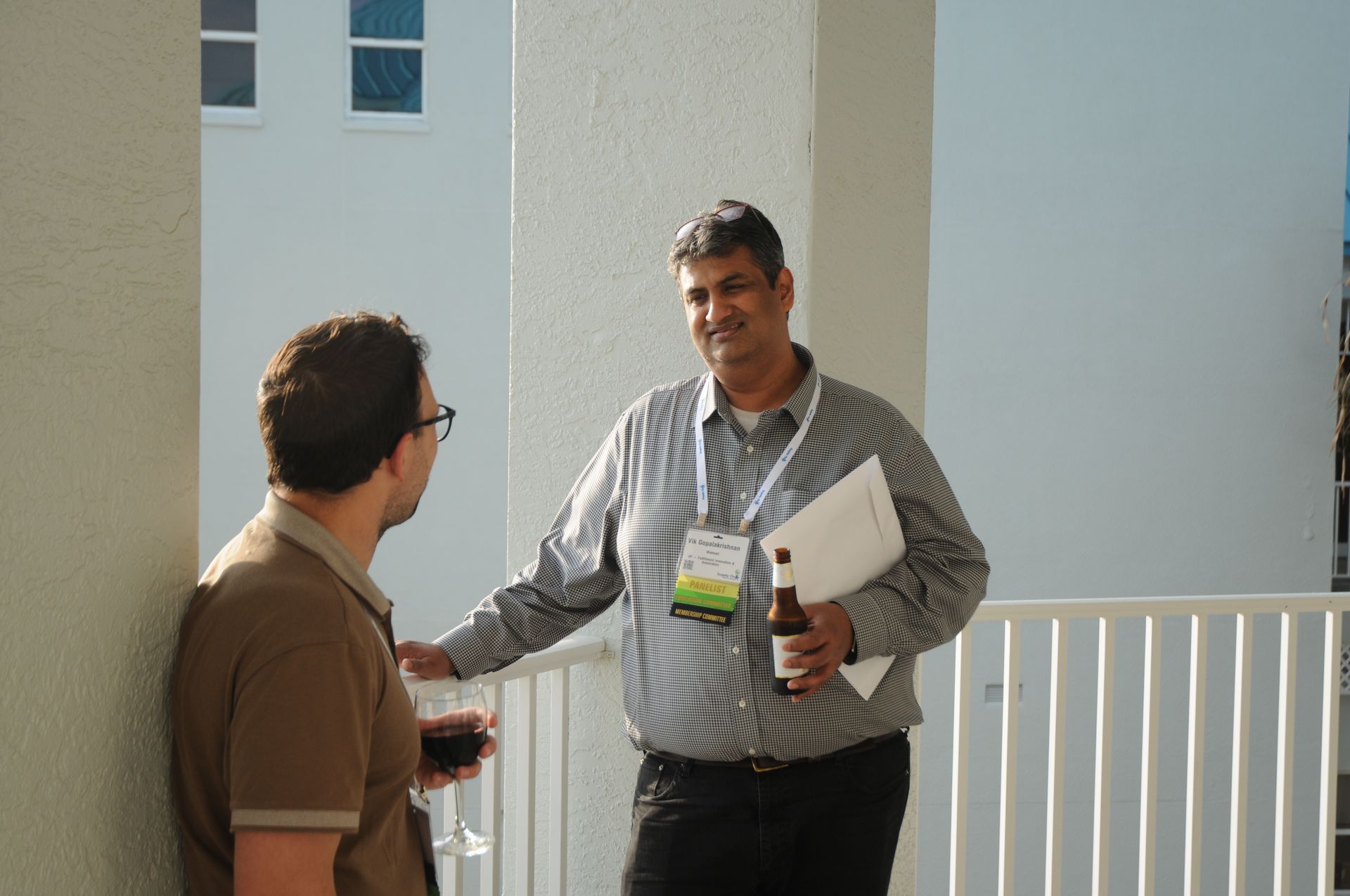 Two men talking on a balcony, one holding a drink and papers, sunlight.