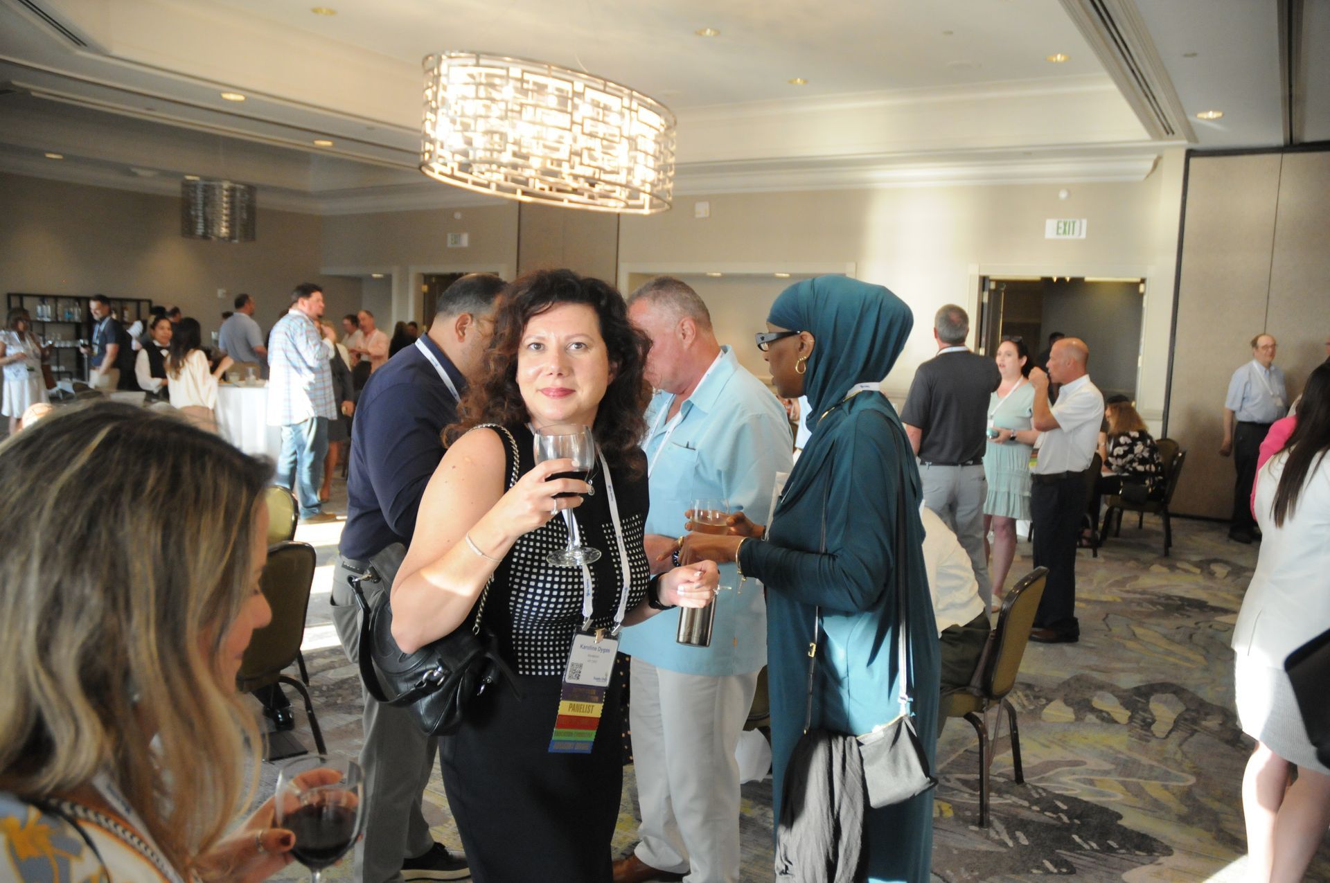 People socializing at an event, woman in foreground holding a drink. Bright, indoor setting.