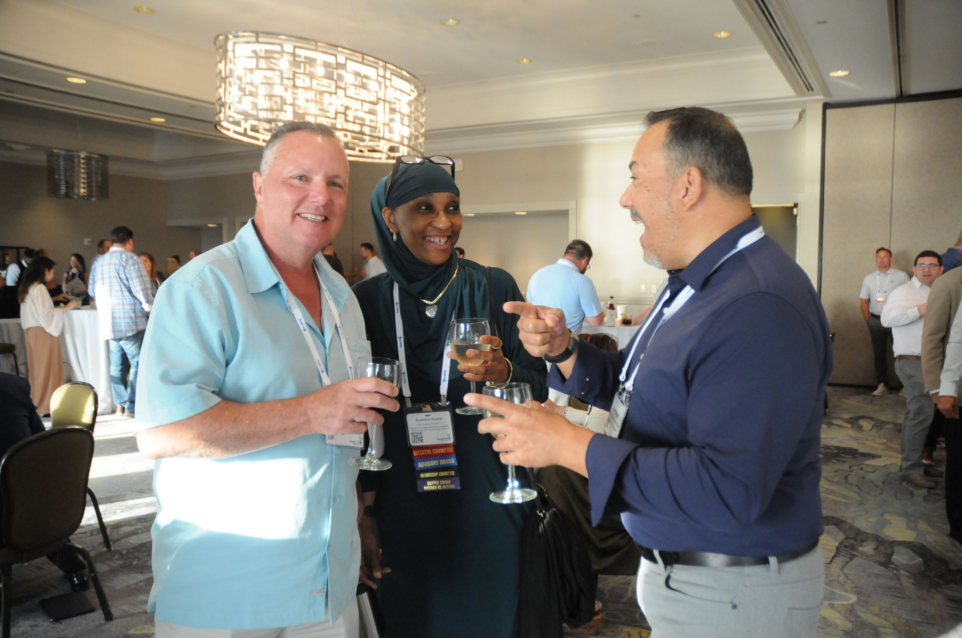Three people at a social gathering; man in blue shirt, woman in headscarf, man laughing. Champagne flutes, large chandelier.