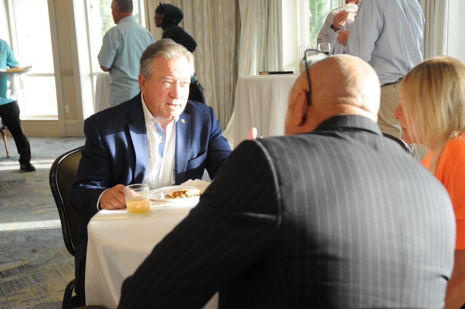 Man in suit seated at a table, conversing with others in a sunlit room.