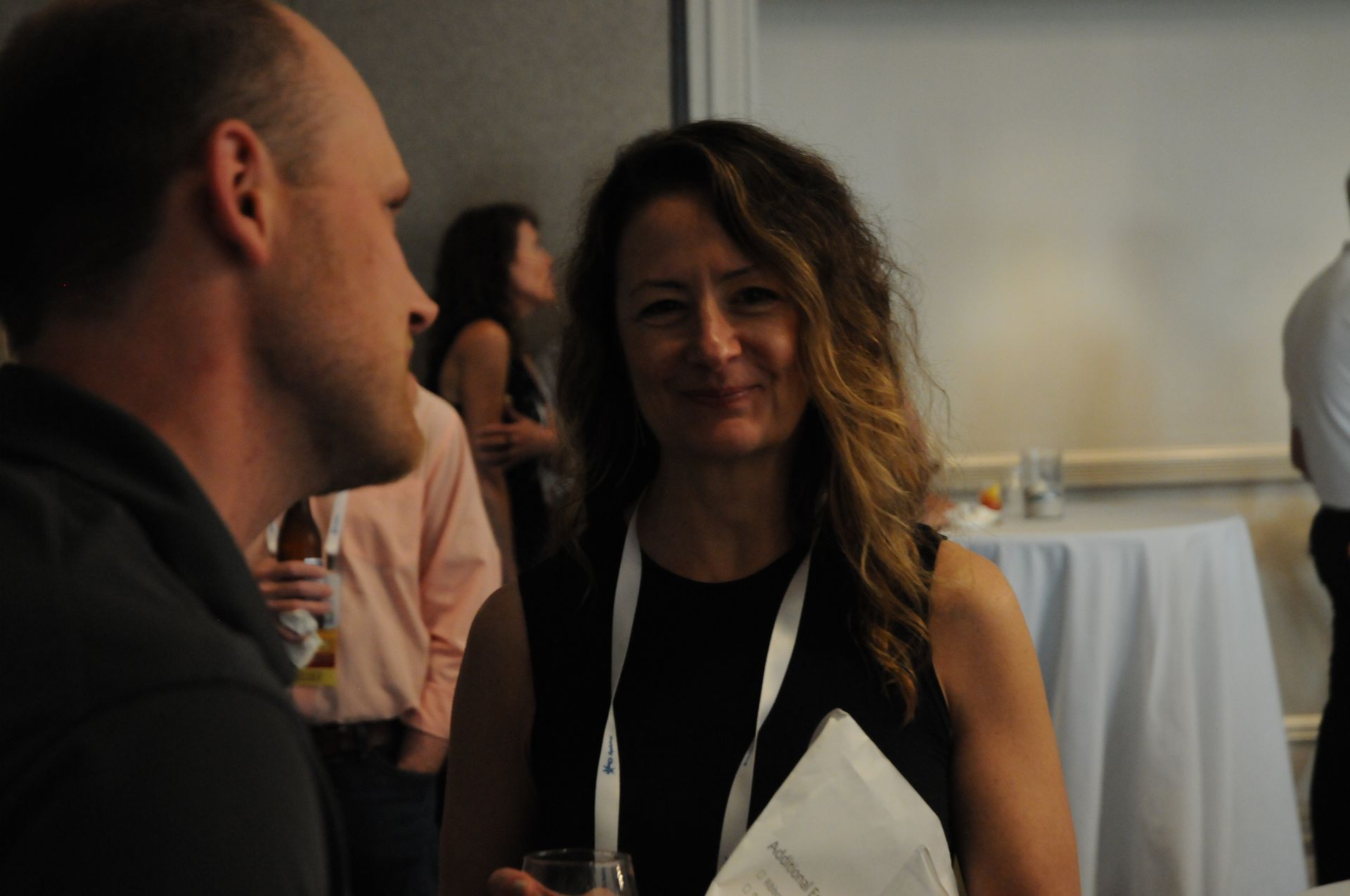 Woman smiling at camera, standing near a man at an event, wearing a black top and lanyard.