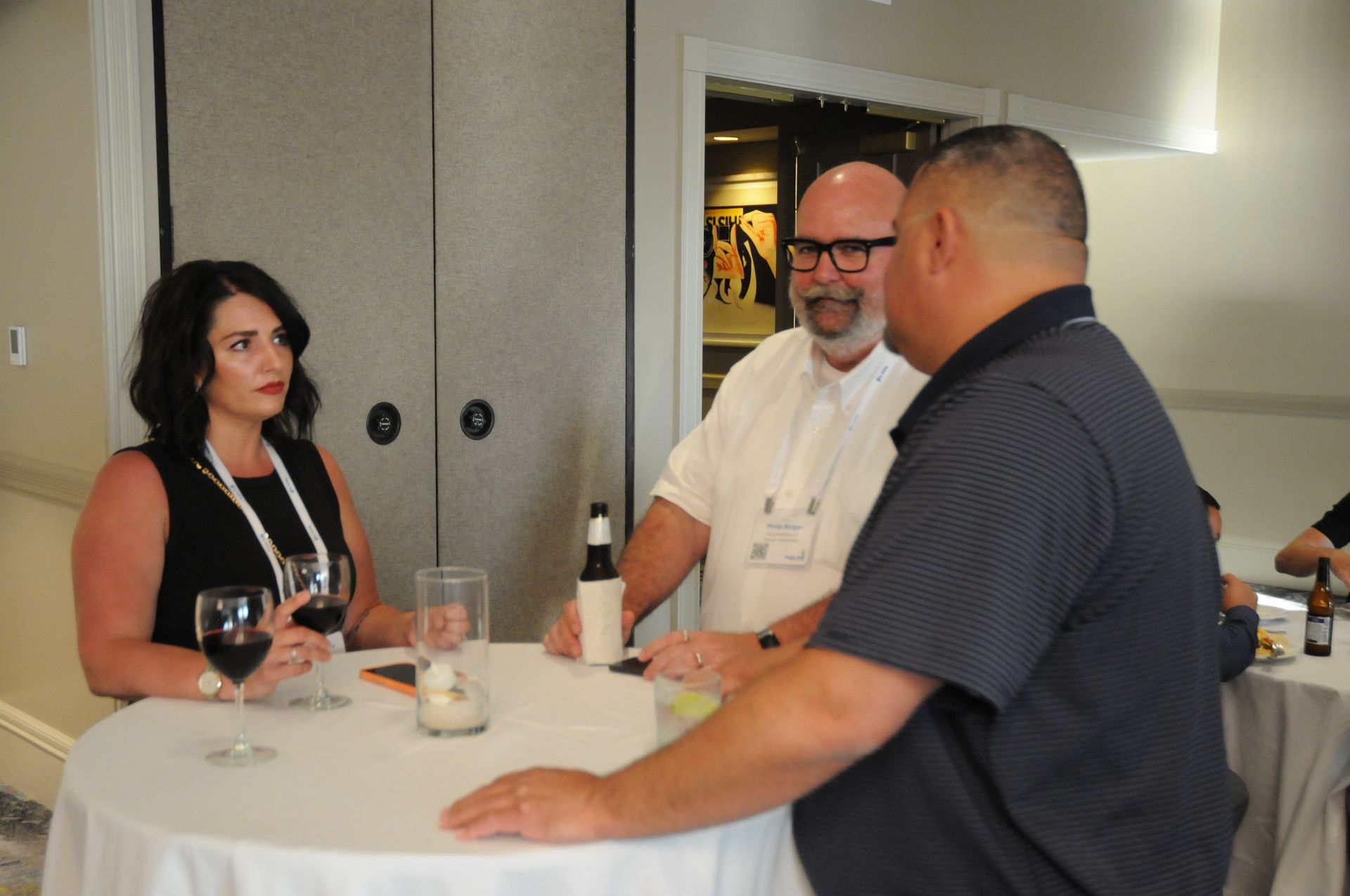 Three people conversing around a small white table in a room, holding drinks.