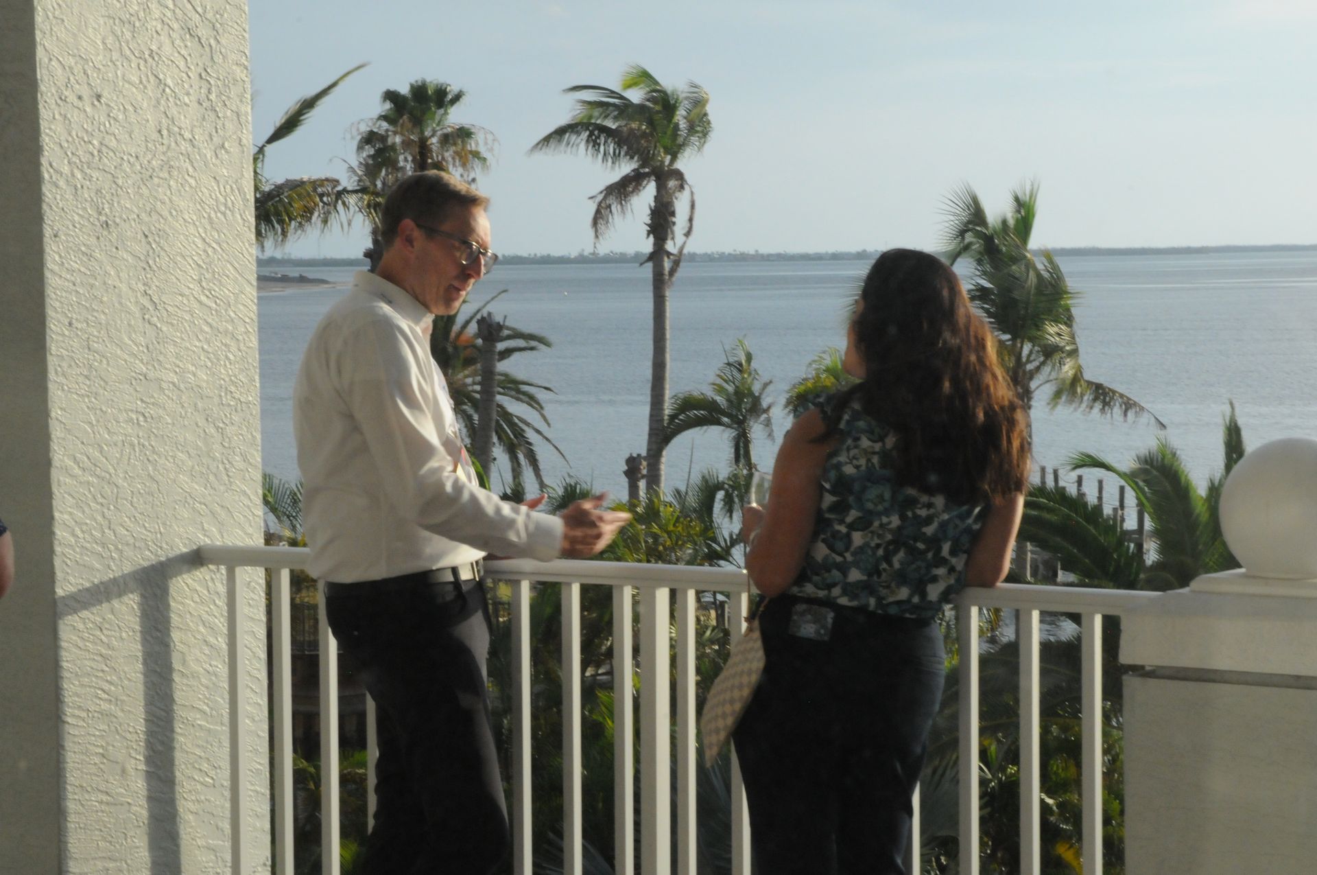 Man and woman on a balcony overlooking the ocean, palm trees visible.