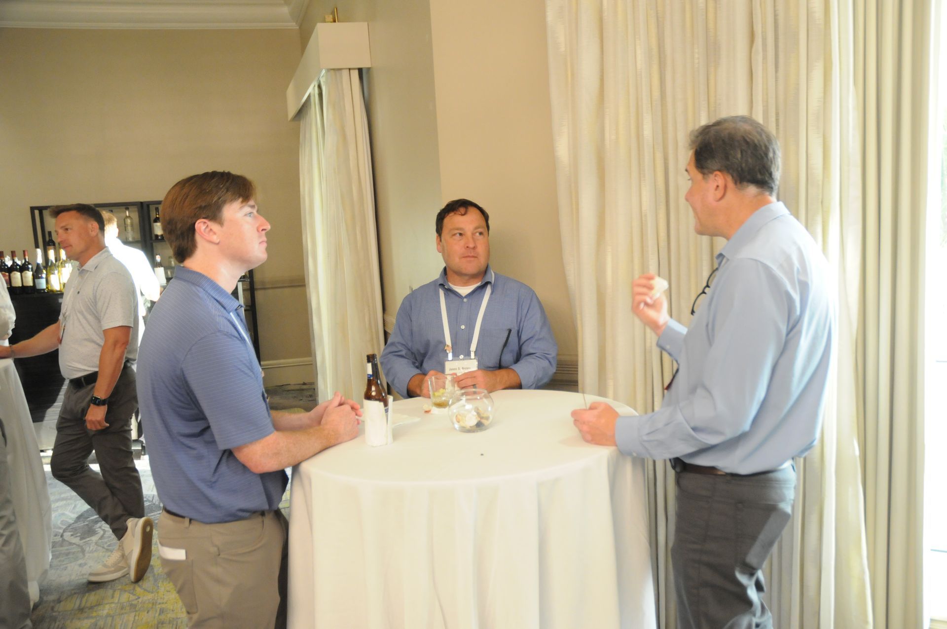 Four men in a bright room converse near a round table; one man gestures as he speaks.