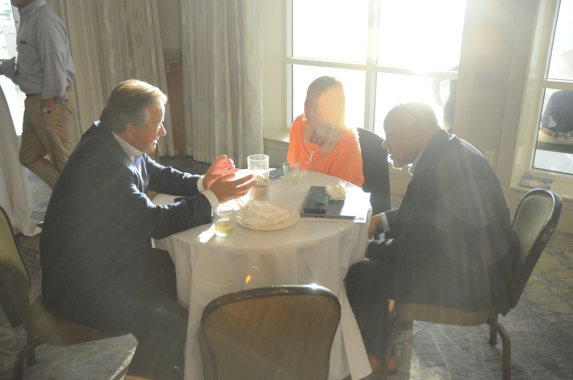 Three people seated around a small round table, conversing in a sunlit room.