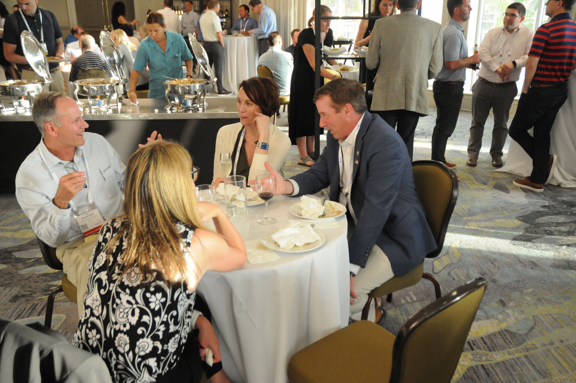 Group of people at a social event: talking around a table with food and drinks, in a bright room.
