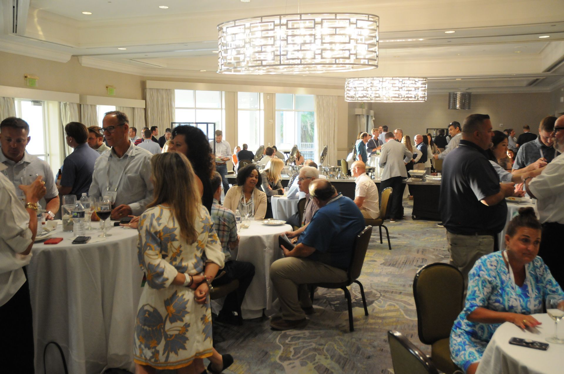 People mingling at an event in a brightly lit ballroom, with round tables and a chandelier.