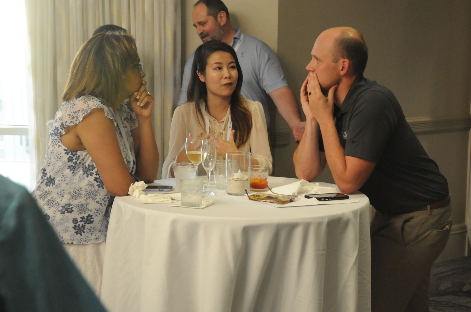 Four people conversing around a small table at a social gathering, some holding drinks, indoor setting.