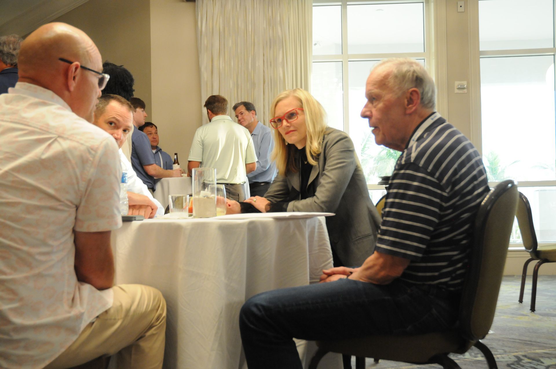 People seated around a table in a room, engaged in conversation. Several others stand in the background.