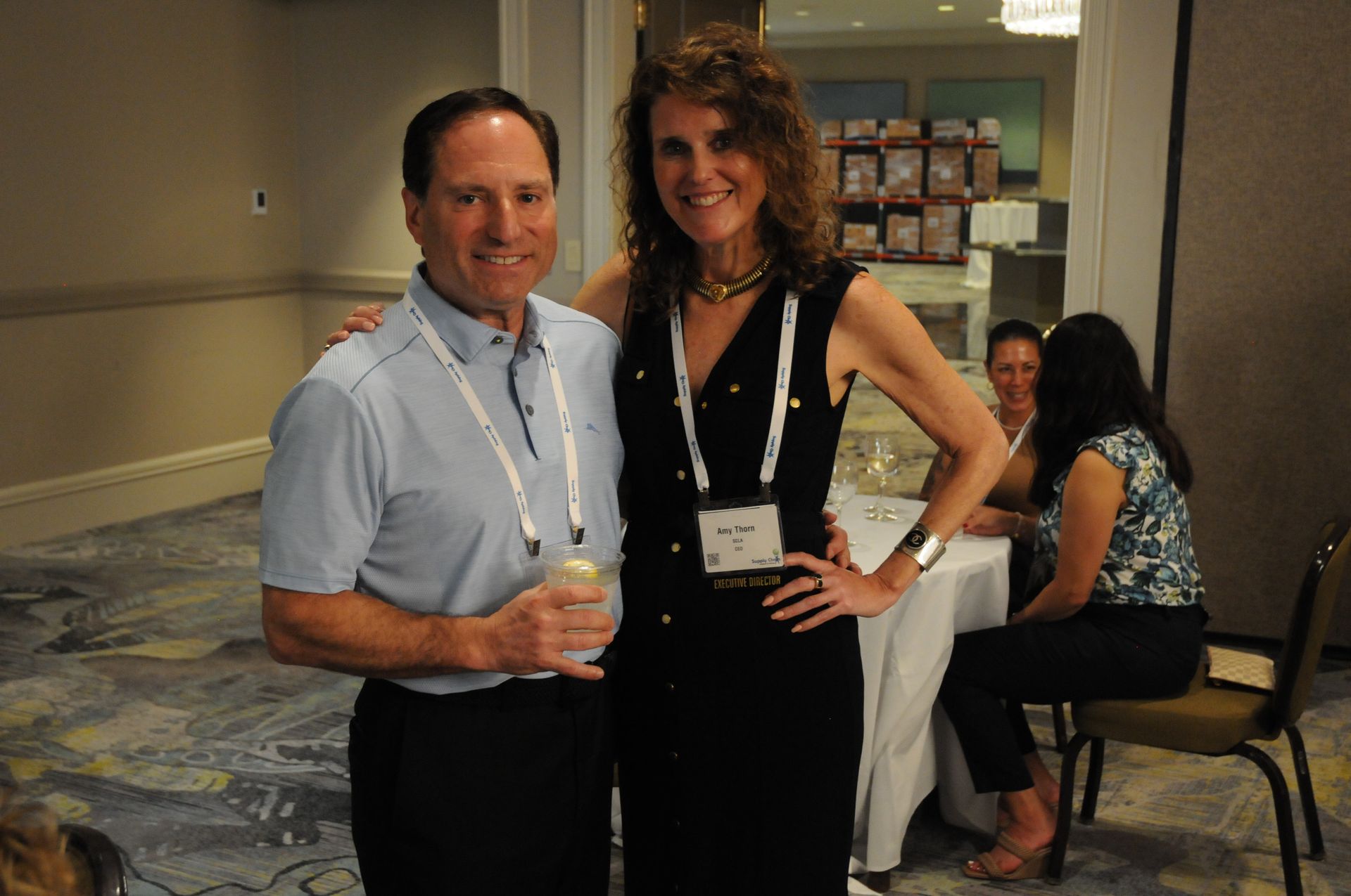 Man and woman pose for photo indoors; both smiling. Man holds drink; woman has hand on hip.