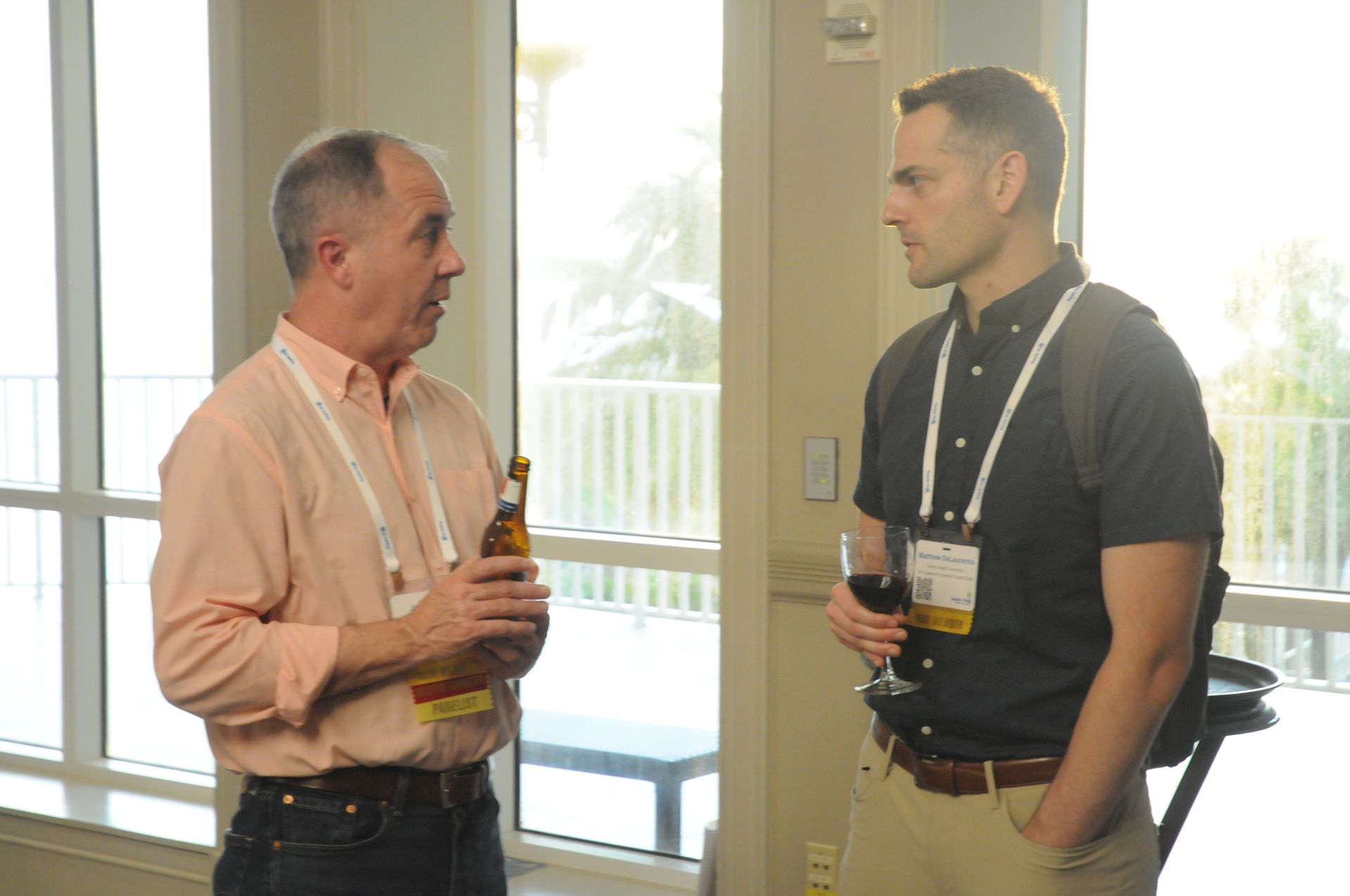 Two men converse near a window, one holds a beer, the other wine; daytime, light-filled room.