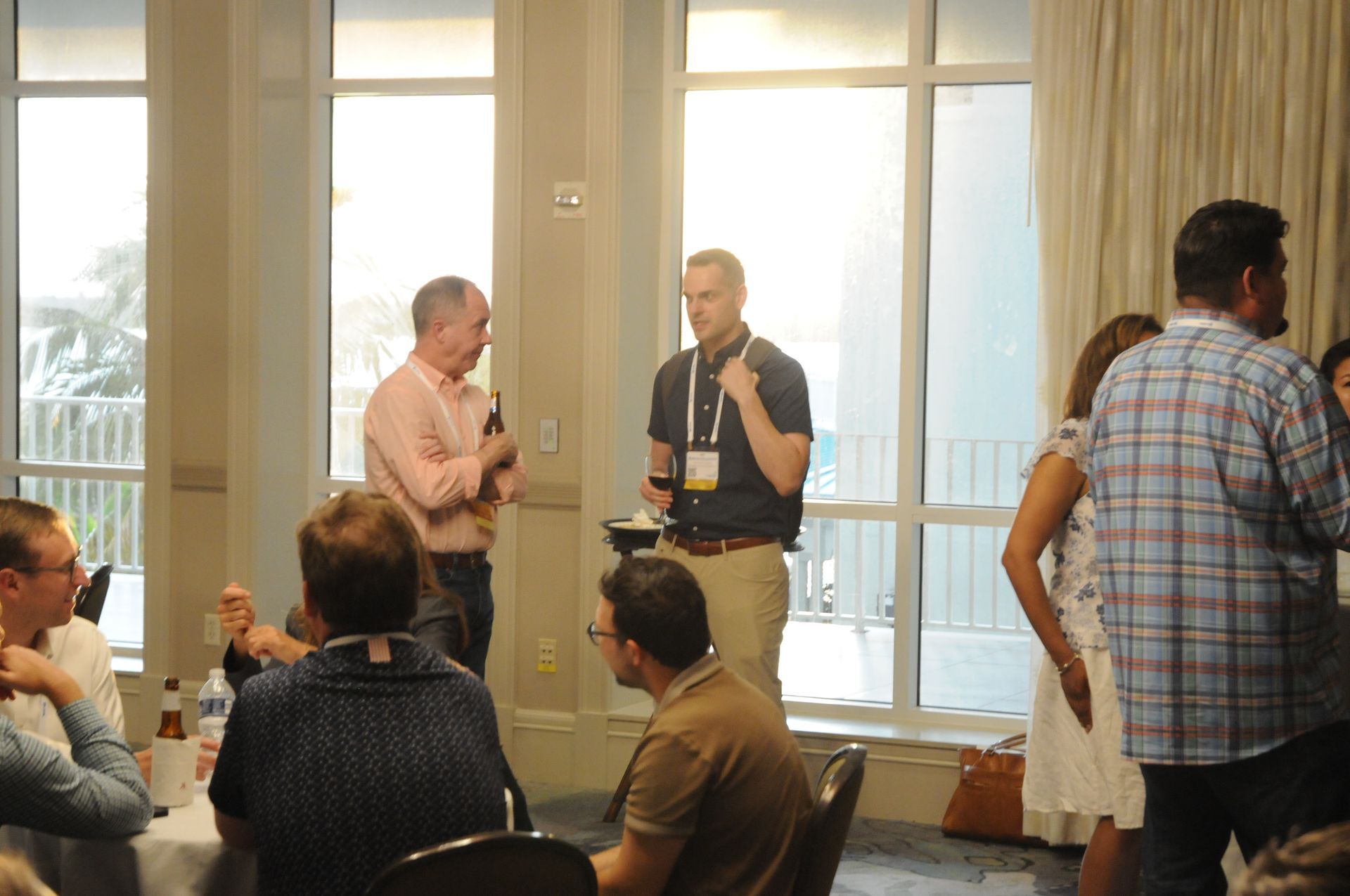 People at a gathering, some talking. One man holds a microphone. Light from windows illuminates the room.