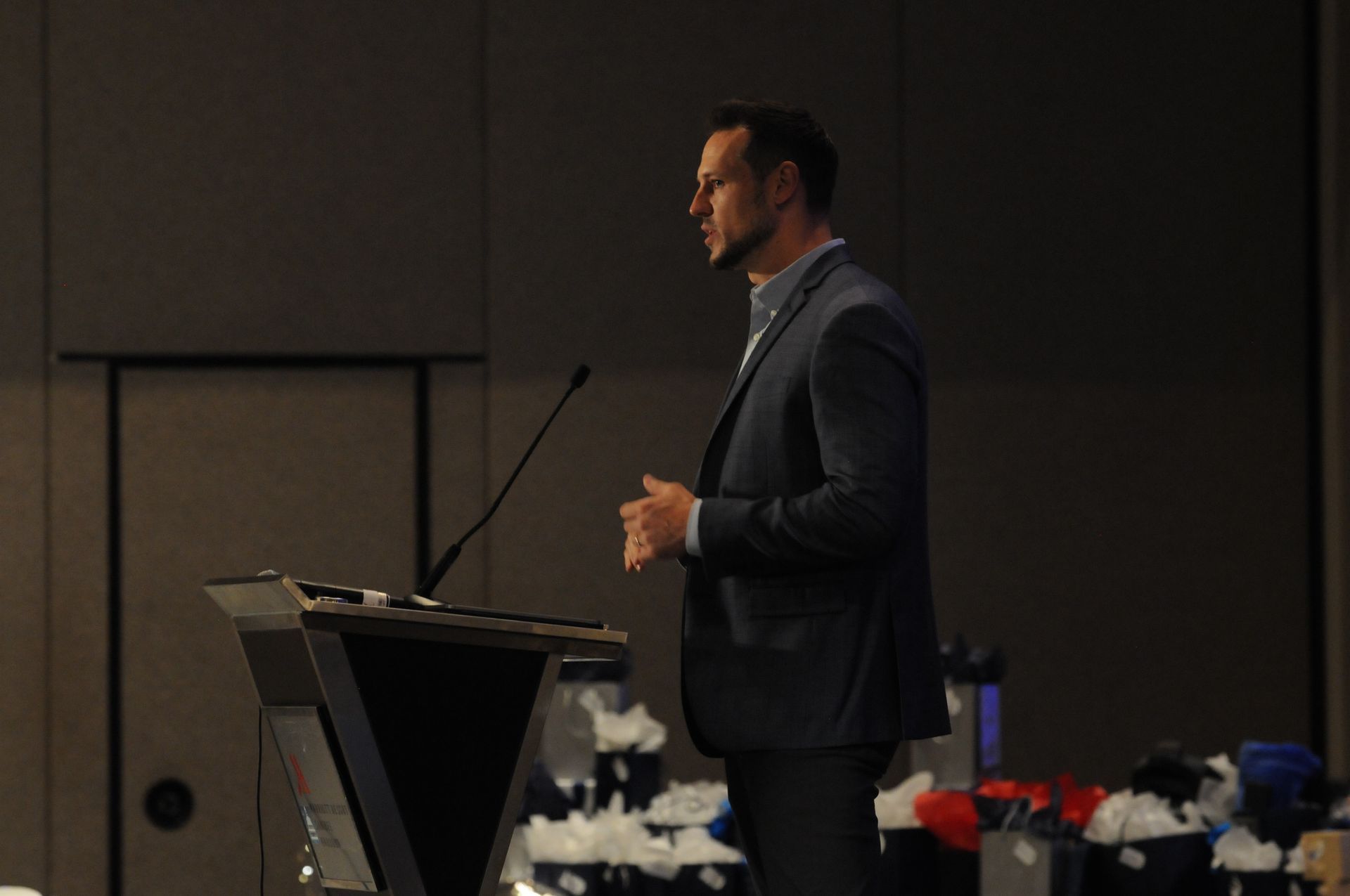 Man in suit speaking at a podium, hands clasped. Dark room with gray walls, microphone.