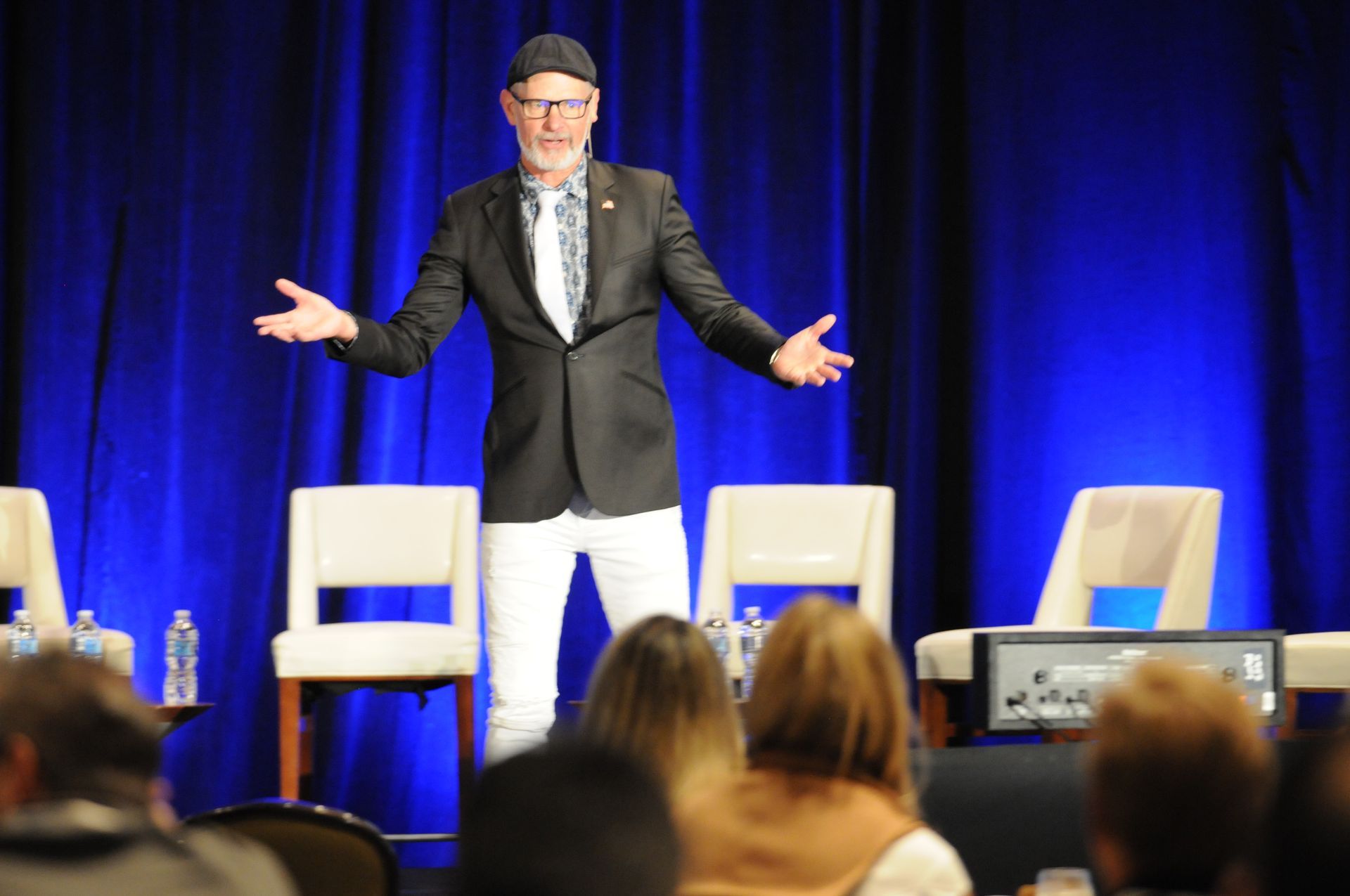Man in black blazer and white pants on stage with arms outstretched, speaking to an audience.