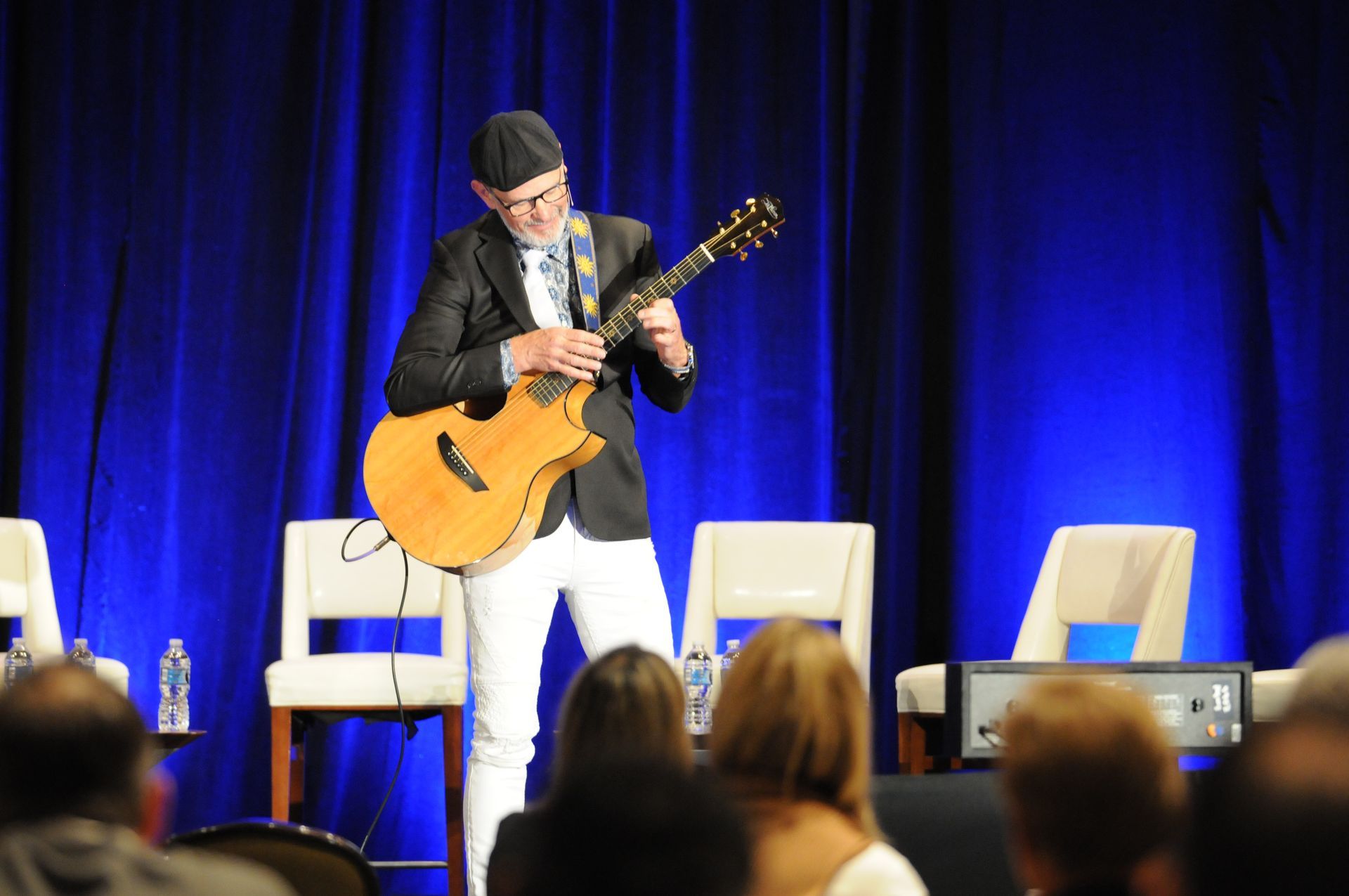 Man in blazer and white pants playing acoustic guitar onstage. Blue curtains and chairs in background.