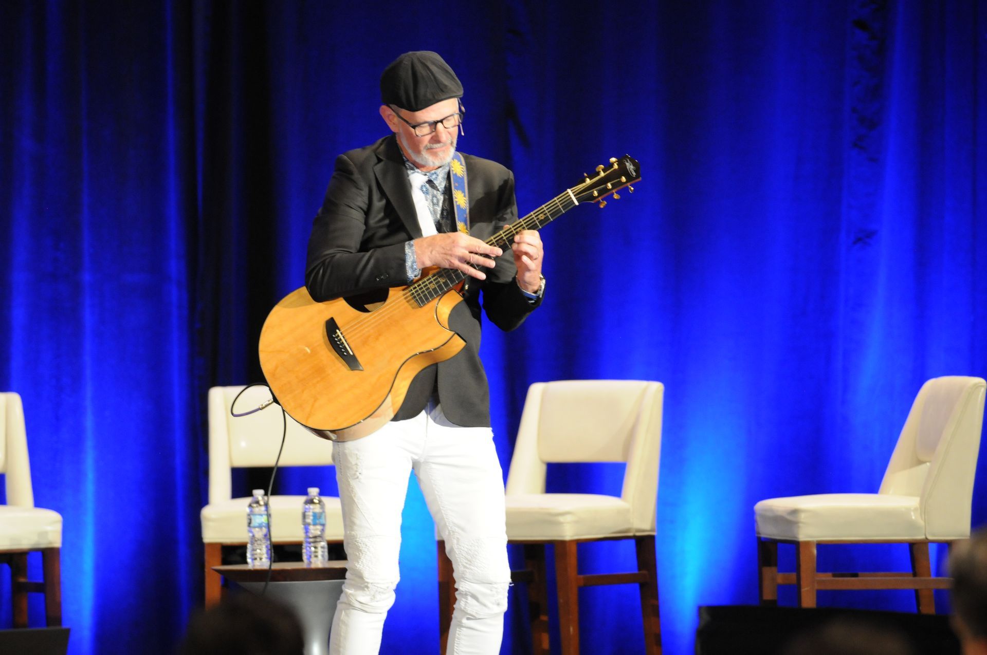 Man in white pants playing guitar on stage, blue curtain backdrop.
