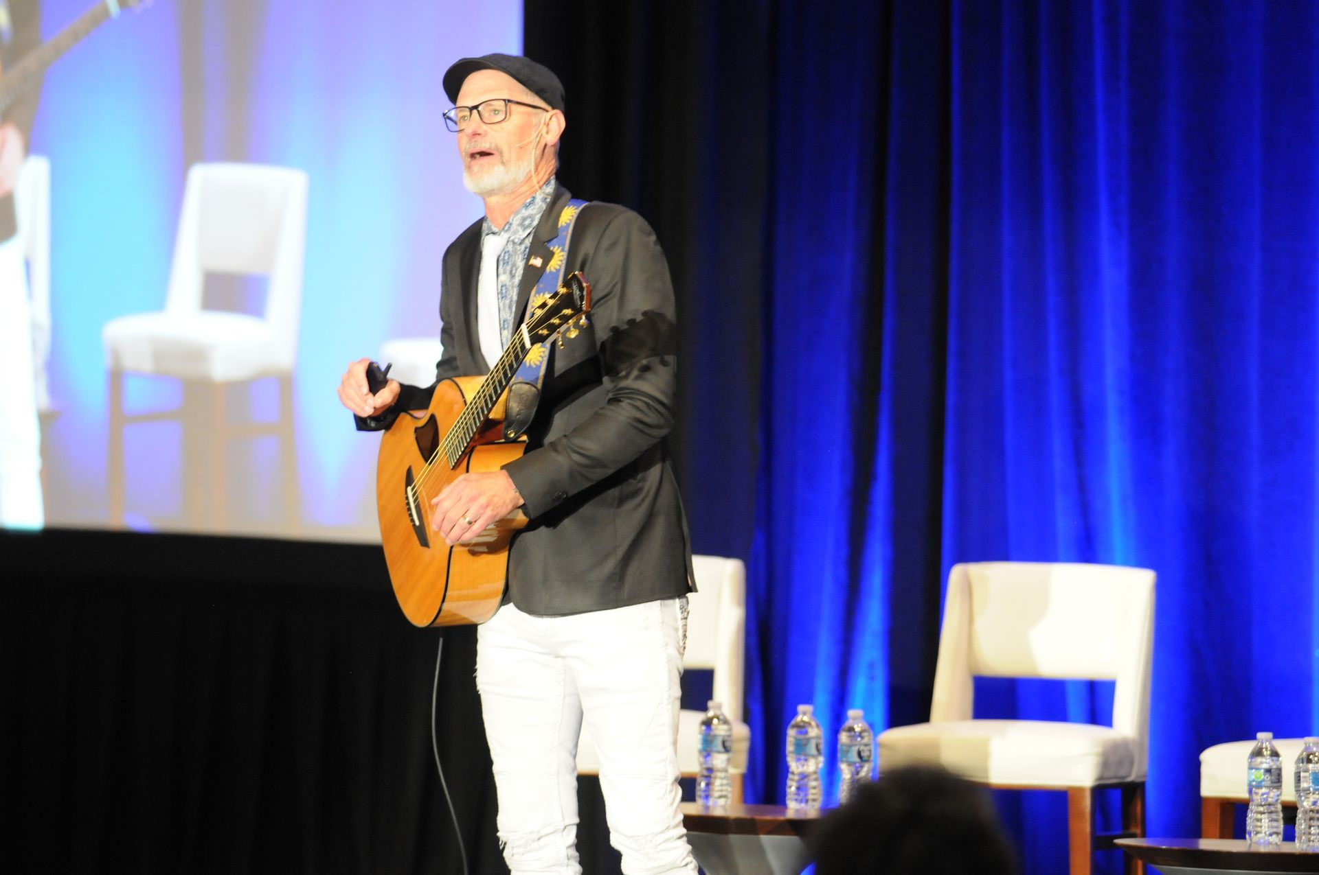 Man in blazer, white pants, and cap singing with acoustic guitar onstage at conference.