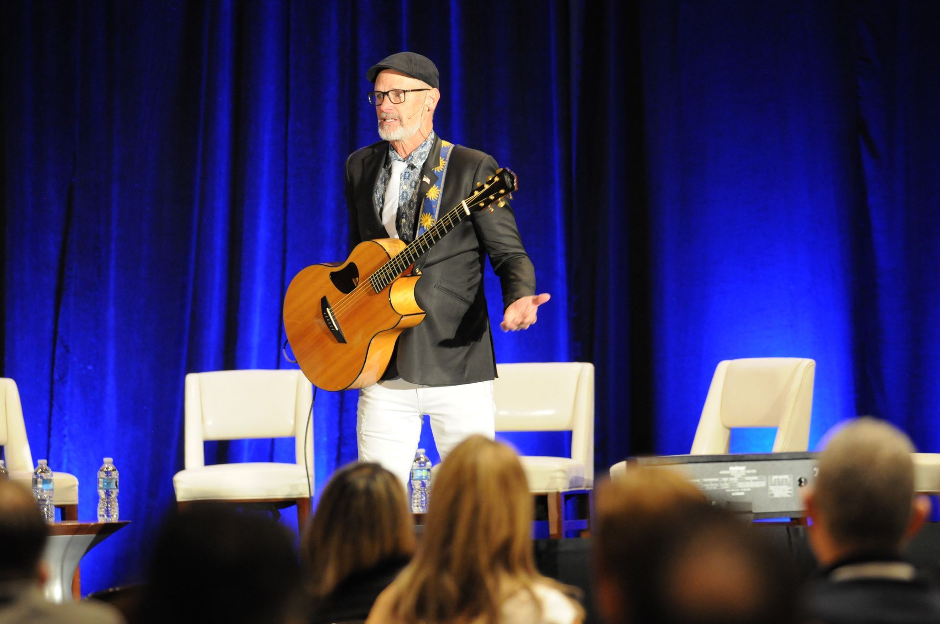 Man on stage with guitar, wearing a black jacket and cap, white pants, blue backdrop, speaking to audience.