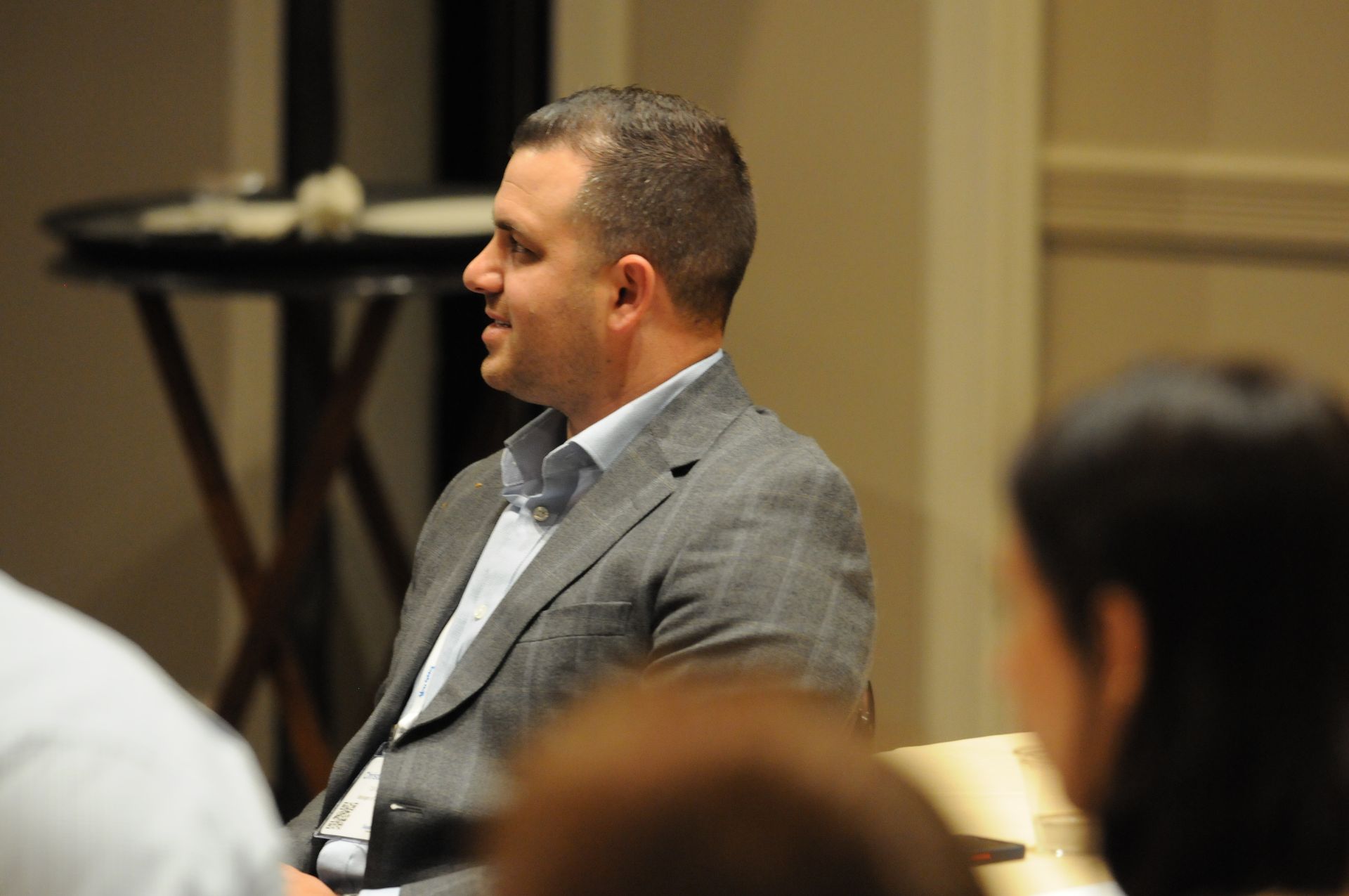 Man in a gray suit jacket smiles at a meeting. Beige-walled room. Others are present.