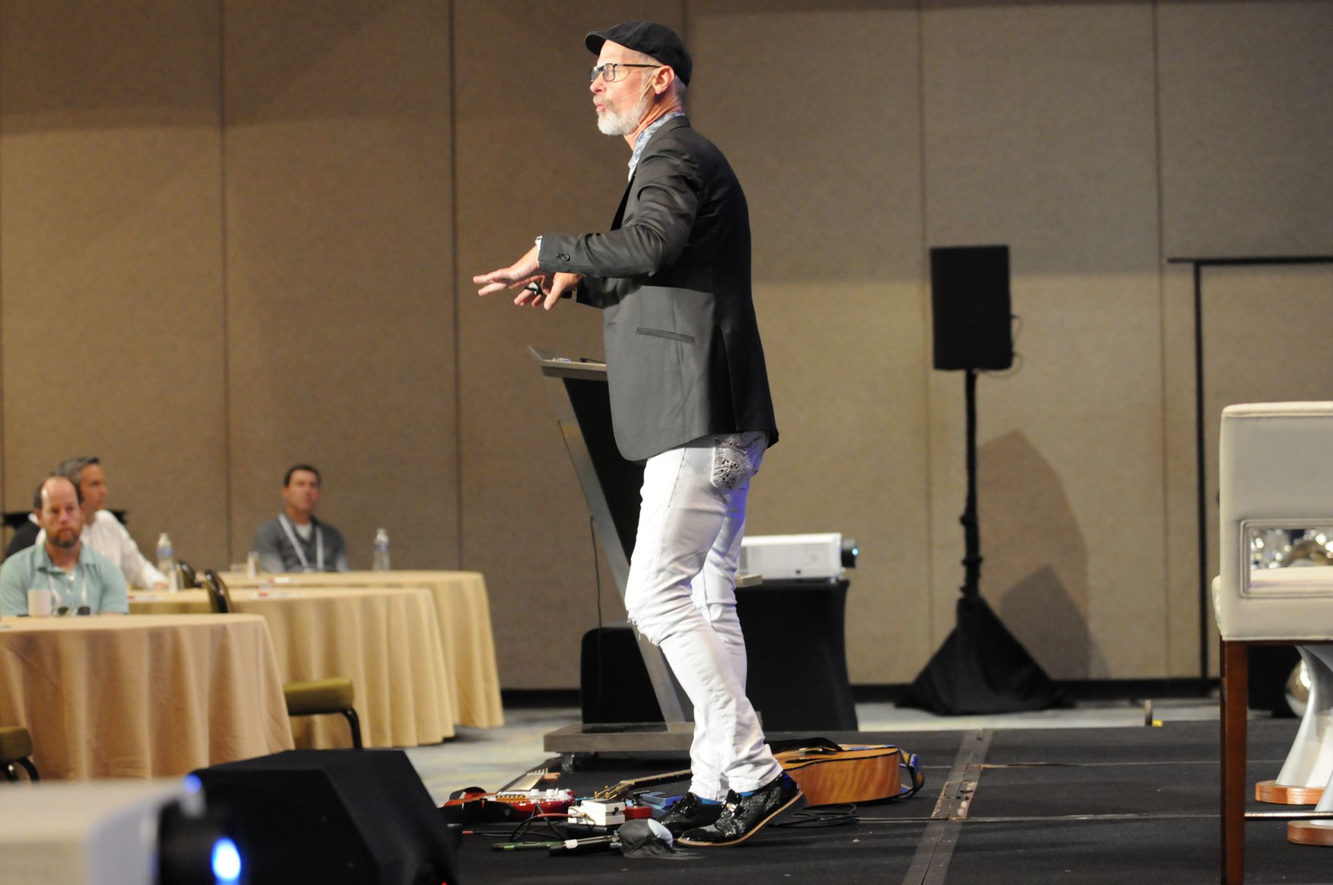 Man in black blazer, white pants speaking on stage, gesturing with hands. Conference room setting.