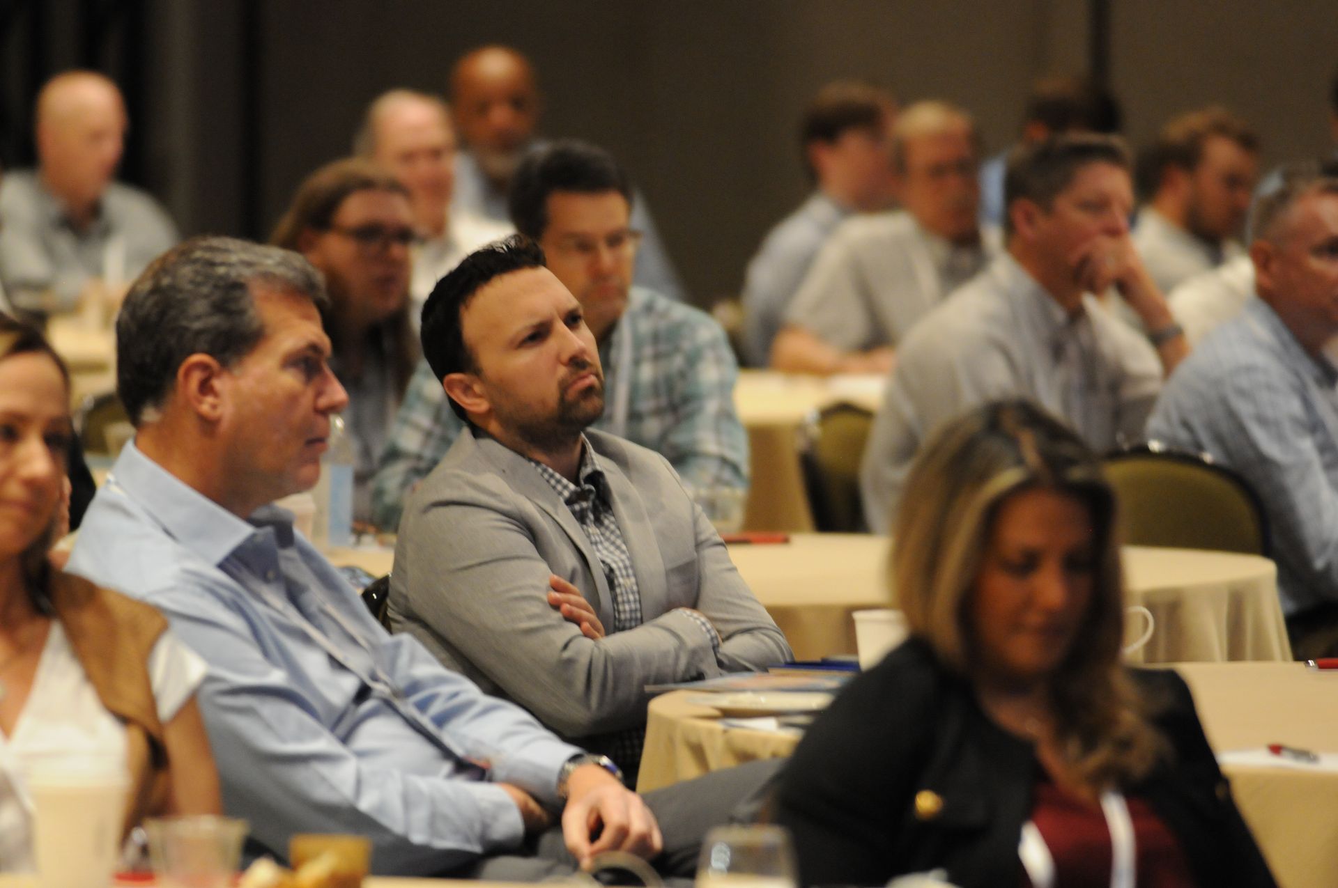 Attendees in a conference room listening attentively. People of various ages and attire are seated at round tables.