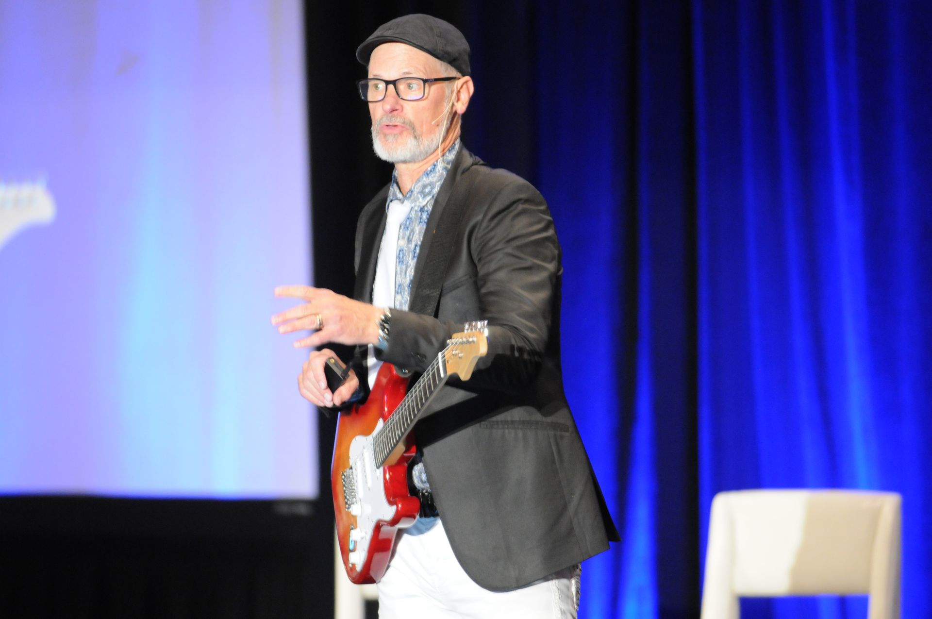 Man in a suit holding a guitar, giving a presentation onstage. Blue backdrop.