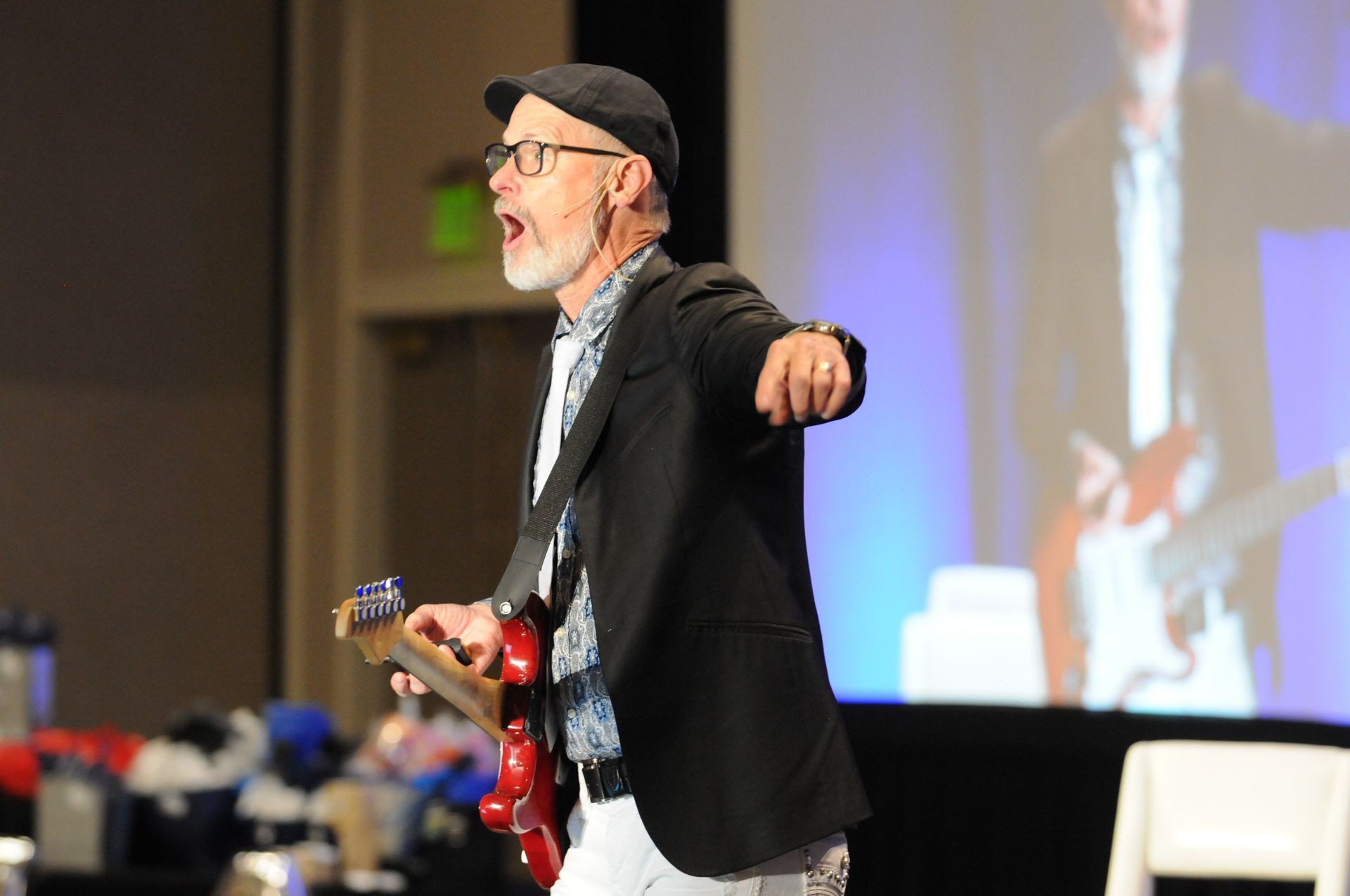 Man on stage plays red guitar, singing, wearing a black jacket, cap, and white pants, in front of a screen with an image.