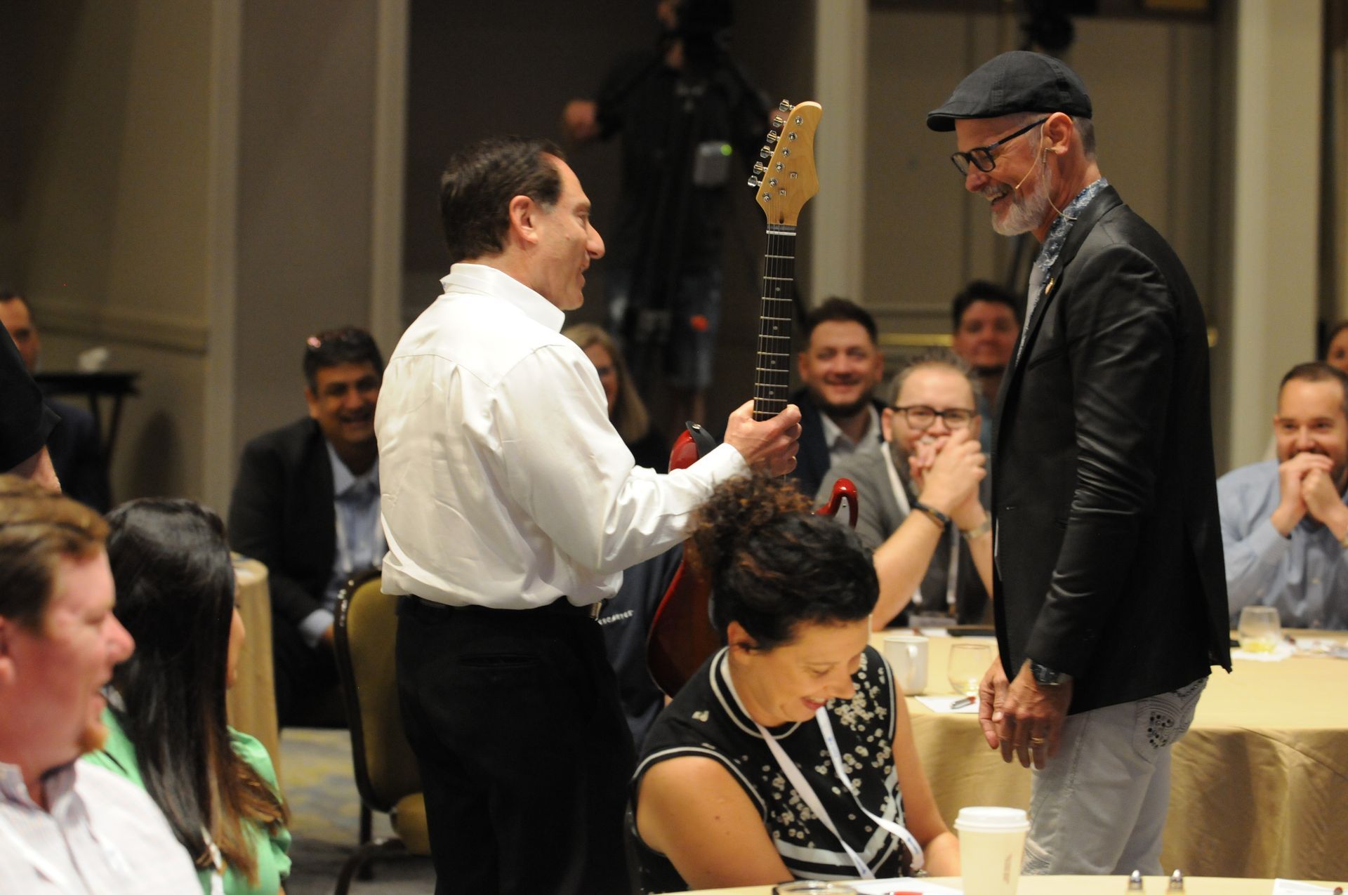Man giving a guitar to another man at a formal event; onlookers smile.