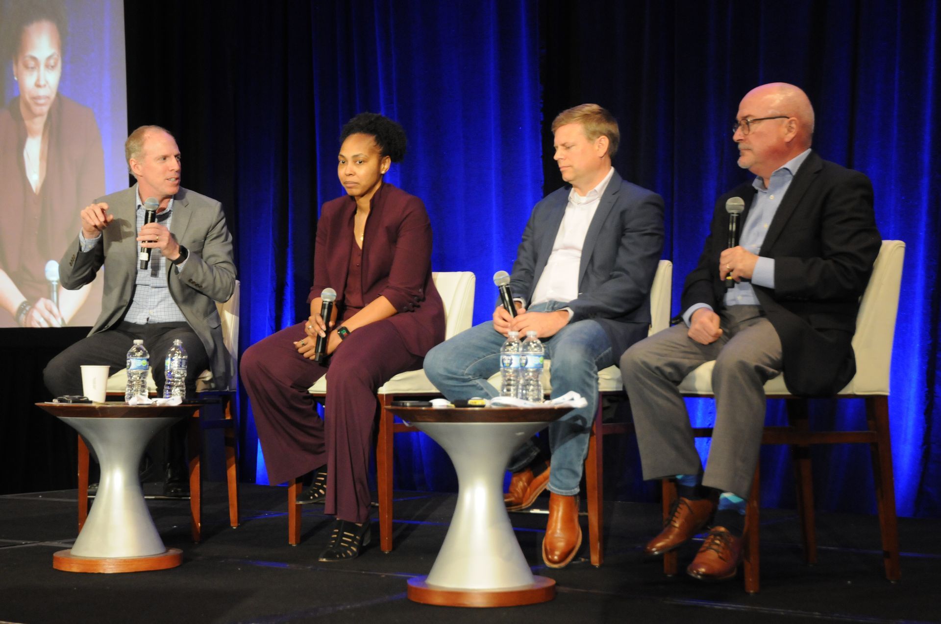 Panel of four people seated at a table, discussing. A screen displays a woman. Blue and purple backdrop.