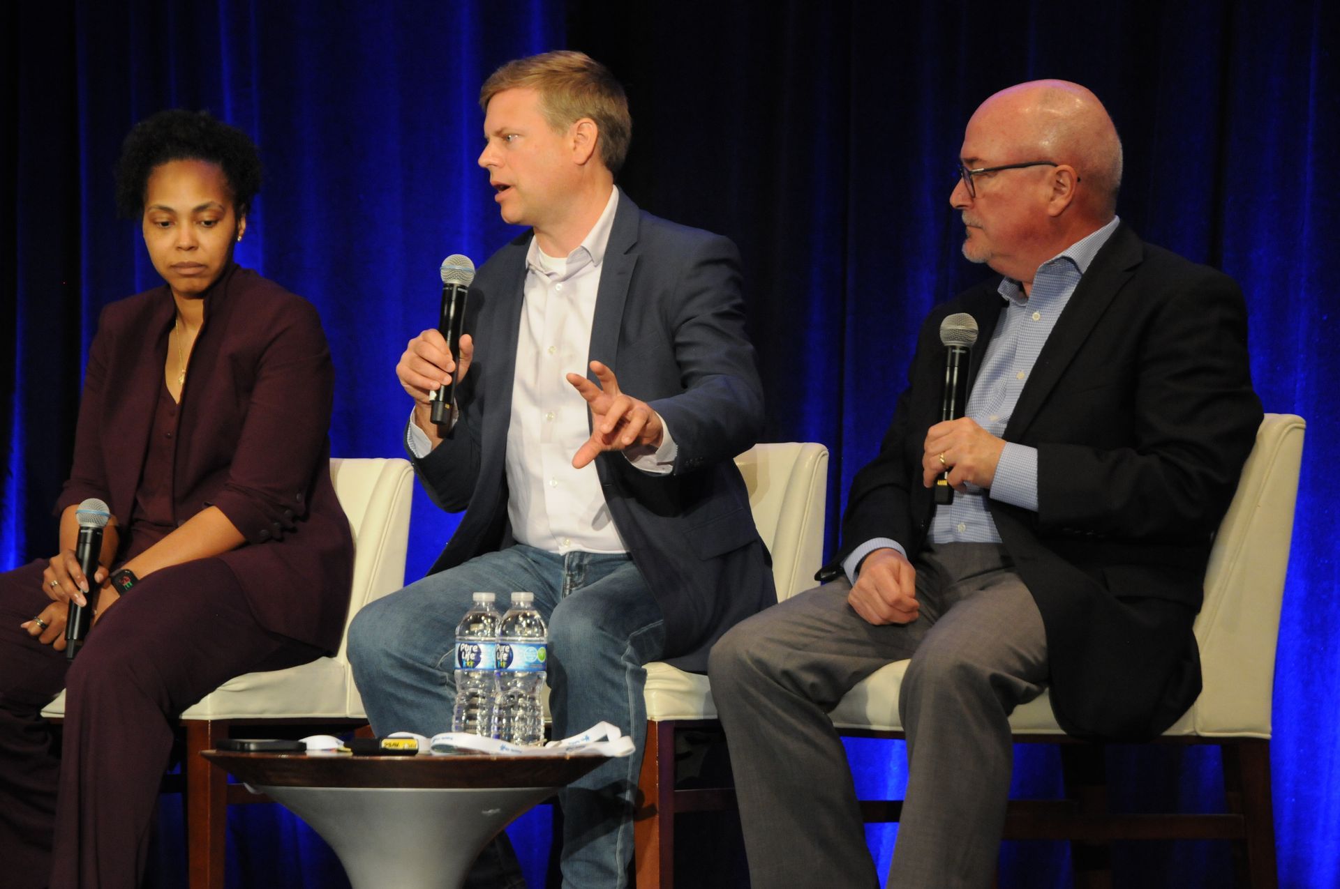 Three panelists on stage with microphones; woman in purple, man in blue blazer, and man in suit jacket.