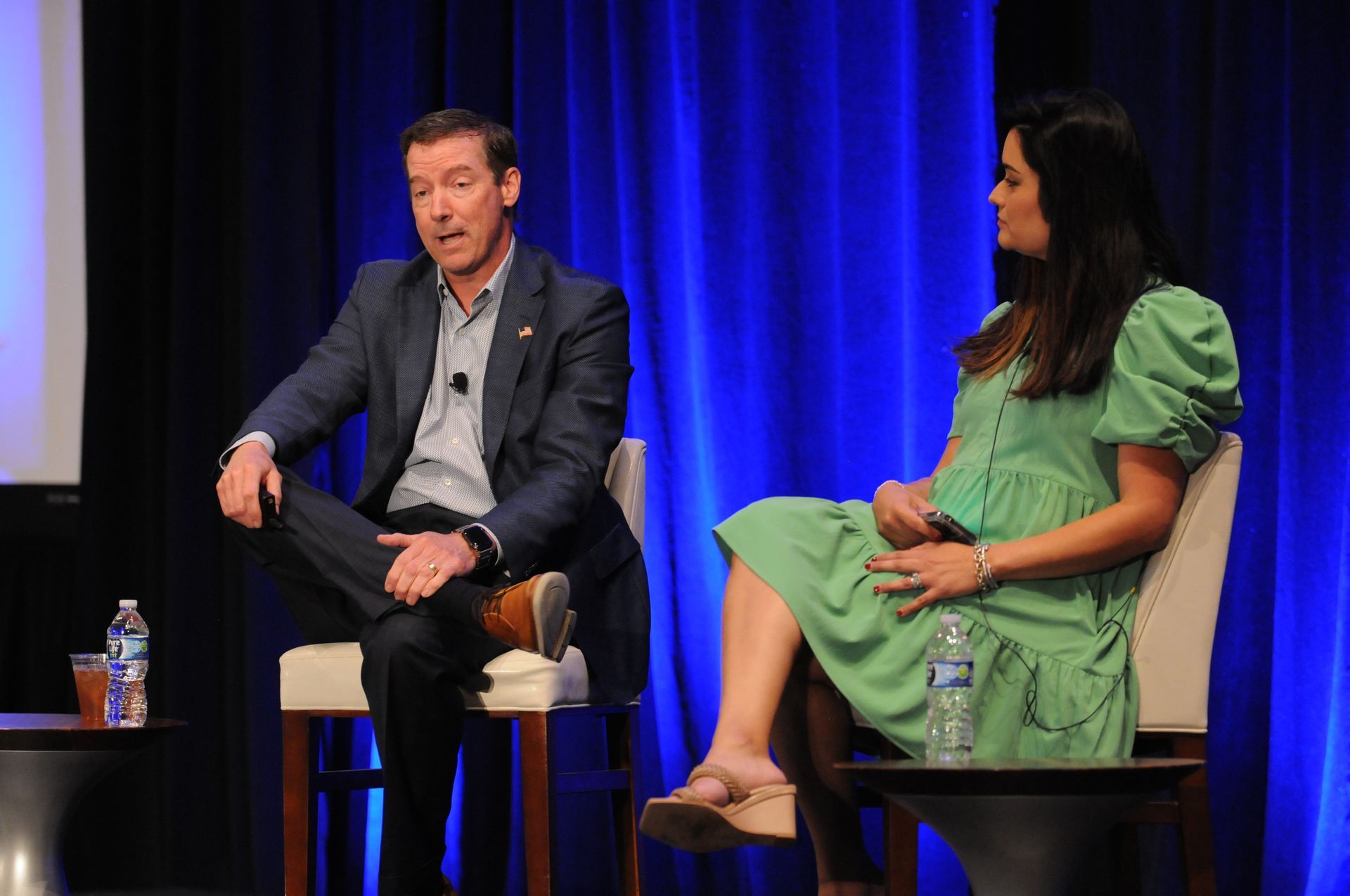 Man and woman on stage, speaking. Man in suit, woman in green dress. Blue curtain backdrop.