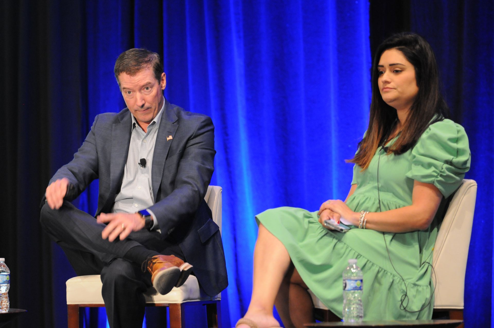 Man and woman seated on stage, facing forward. Man in blazer, woman in green dress. Blue backdrop.