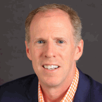 Smiling man with fair skin, in a blue blazer, orange checked shirt, against a gray backdrop.