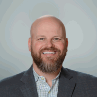 Bald man with a beard smiles broadly, wearing a grey suit jacket over a blue checkered shirt.