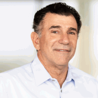 Man in a light blue shirt smiling, looking at the viewer; indoor, neutral background.