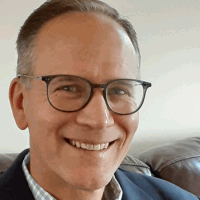 Man with glasses smiles, wearing a blazer, sitting inside.