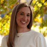 Woman with long blonde hair smiles in front of a yellow and green tree. She wears a cream sweater.