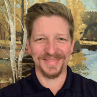 Man with light brown hair and beard smiles in front of a nature painting, wearing a dark blue shirt.