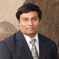 Man in a suit and tie, smiling slightly, standing outdoors with a rock background.