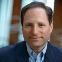 Man in blue blazer and shirt, smiling slightly. Blurred background.