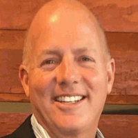 Bald man with a friendly smile wearing a blazer. He is indoors against a wooden backdrop.