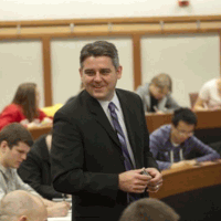 Professor in a suit, smiling in a classroom. Students in the background, seated.