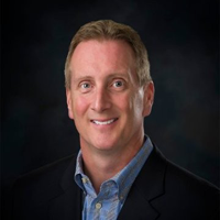 Middle-aged man with blonde hair smiles, wearing a blue shirt and navy blazer, dark background.