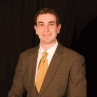 Young man in a brown suit and gold tie smiles at the camera. Black background.
