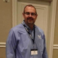 Man wearing glasses, blue shirt, and lanyard, smiling at event.